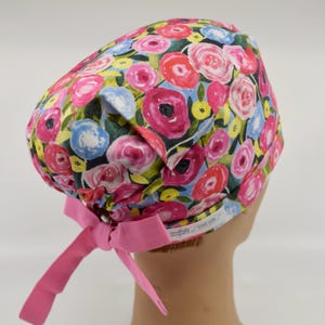 May include: A floral patterned scrub cap featuring a vibrant watercolor design of pink, red, blue, and yellow flowers on a black background. The cap has a pink bow at the back. The cap is designed for medical professionals.