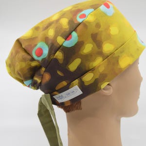 May include: A patterned scrub cap featuring a design reminiscent of a brown trout. The cap has a yellow, brown, and blue pattern, with a green tie at the back. A small white tag is sewn on the side.