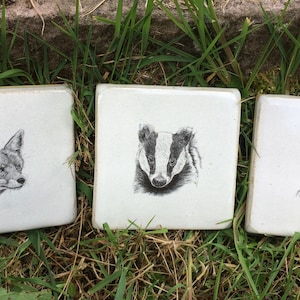 May include: Three white ceramic tiles with black and white illustrations of a fox, a badger, and a rabbit.