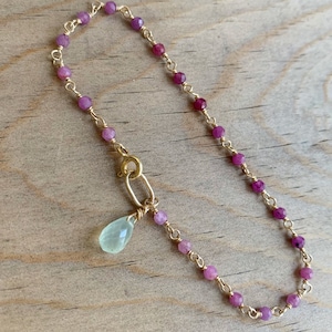Pode incluir: Um delicado bracelete de corrente dourada com pequenas contas rosa e roxas e um pingente de pedra preciosa verde claro.