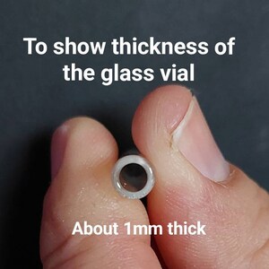 May include: A close-up of a glass vial held between two fingers, showcasing its thickness. The text reads, "To show thickness of the glass vial" and "About 1mm thick."
