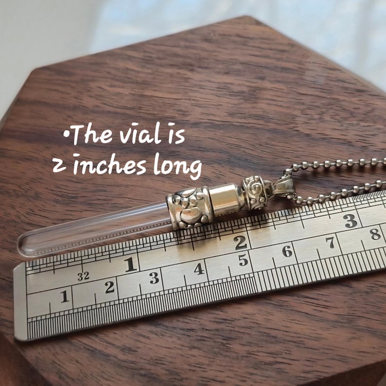 May include: A clear glass vial pendant with silver-colored metal accents and a ball chain. The vial is 2 inches long. The pendant rests on a wooden surface next to a ruler.