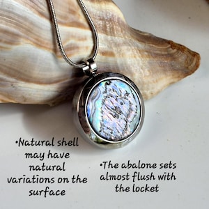 May include: A round silver-toned locket pendant featuring an iridescent abalone shell inlay on a thin snake chain. Text reads: Natural shell may have natural variations on the surface. The abalone sets almost flush with the locket.