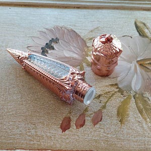 May include: A rose gold lip gloss tube with a decorative, ornate design. The tube is clear and shows the lip gloss inside. The cap is also rose gold and has a decorative design.