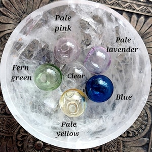 May include: A collection of glass spheres in various colors, including pale pink, fern green, clear, pale lavender, blue, and pale yellow. The spheres are arranged in a clear, textured bowl. Text labels identify each color.