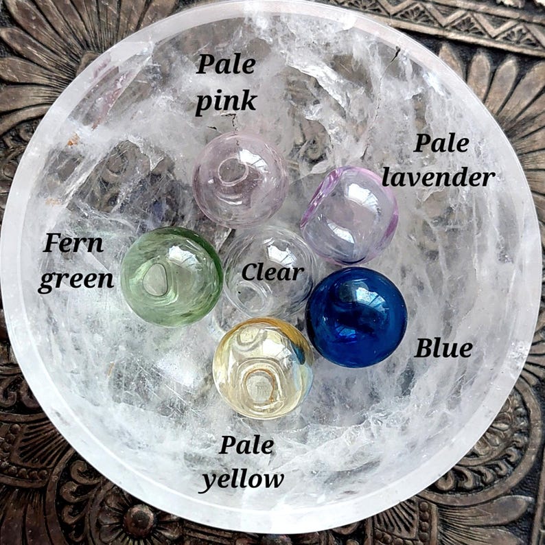 May include: A collection of glass spheres in various colors, including pale pink, fern green, clear, pale lavender, blue, and pale yellow. The spheres are arranged in a clear, textured bowl. Text labels identify each color.