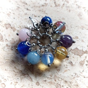 Puede incluir: Un brazalete con charms con una variedad de cuentas de piedras preciosas de colores, incluyendo transparentes, rosas, azules, moradas y amarillas. Las cuentas est&aacute;n unidas a una cadena de plata con detalles met&aacute;licos decorativos.