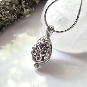 Peut inclure: Un pendentif en métal argenté présentant un design complexe de cage en filigrane ajouré en forme de goutte. Le pendentif est suspendu à un collier fin et flexible en chaîne serpent argentée, avec une finition polie et des volutes détaillées.