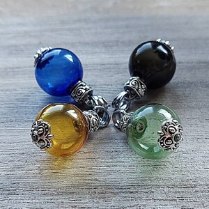May include: Four glass orb pendants in blue, black, yellow, and green. Each orb has a silver-colored decorative cap and a ring for hanging. The pendants are displayed on a light gray wooden surface.