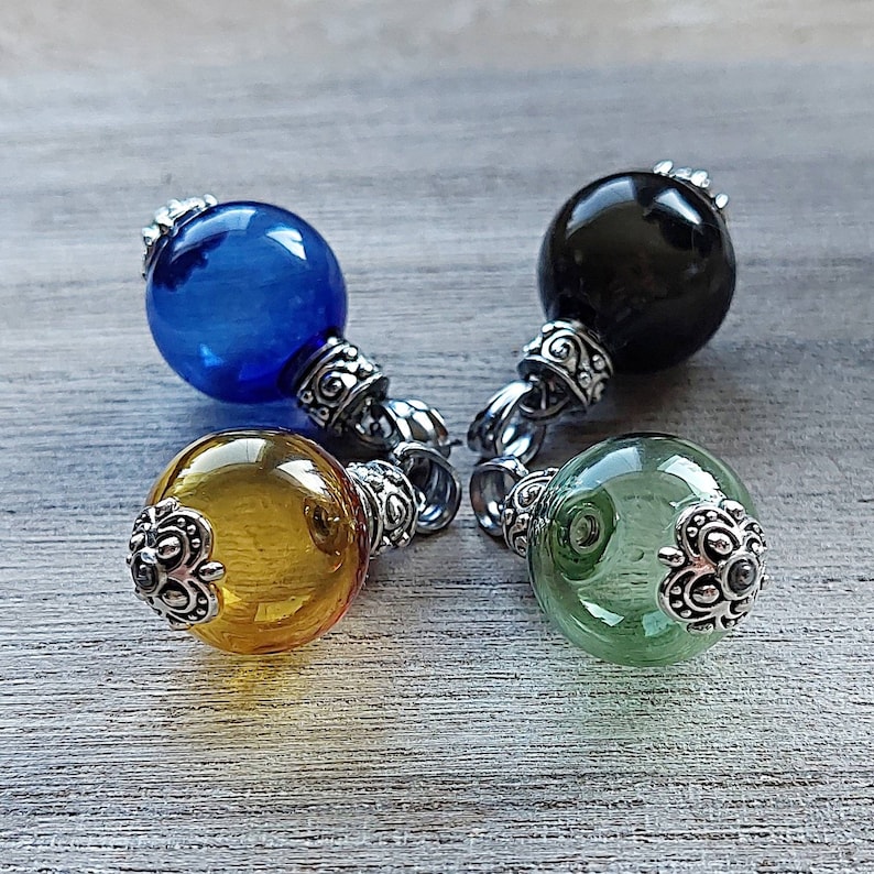 May include: Four glass orb pendants in blue, black, yellow, and green. Each orb has a silver-colored decorative cap and a ring for hanging. The pendants are displayed on a light gray wooden surface.