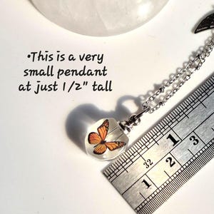 May include: A small pendant featuring an orange and black butterfly encased in clear material, attached to a silver-toned chain. A metal ruler shows the pendant is 1/2 inch tall. Text on the image reads: This is a very small pendant at just 1/2" tall.