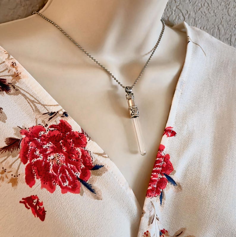 May include: A silver-toned necklace with a clear, cylindrical pendant. The pendant has decorative metal accents and a small, light pink crystal. The necklace is displayed on a mannequin wearing a floral print top.