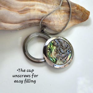 May include: A silver-toned metal locket necklace featuring a circular pendant with an iridescent abalone shell inlay. The locket has a screw-off cap for filling. Text on the image reads: The cap unscrews for easy filling. The chain is a thin snake style.