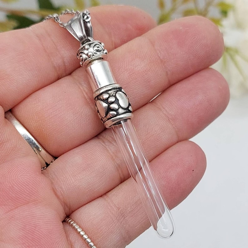 May include: A silver-colored pendant necklace with a clear glass vial. The pendant has decorative metalwork with paw prints and a decorative top. The vial is designed to hold a small amount of cremated remains or other keepsakes. The necklace is displayed on a hand.