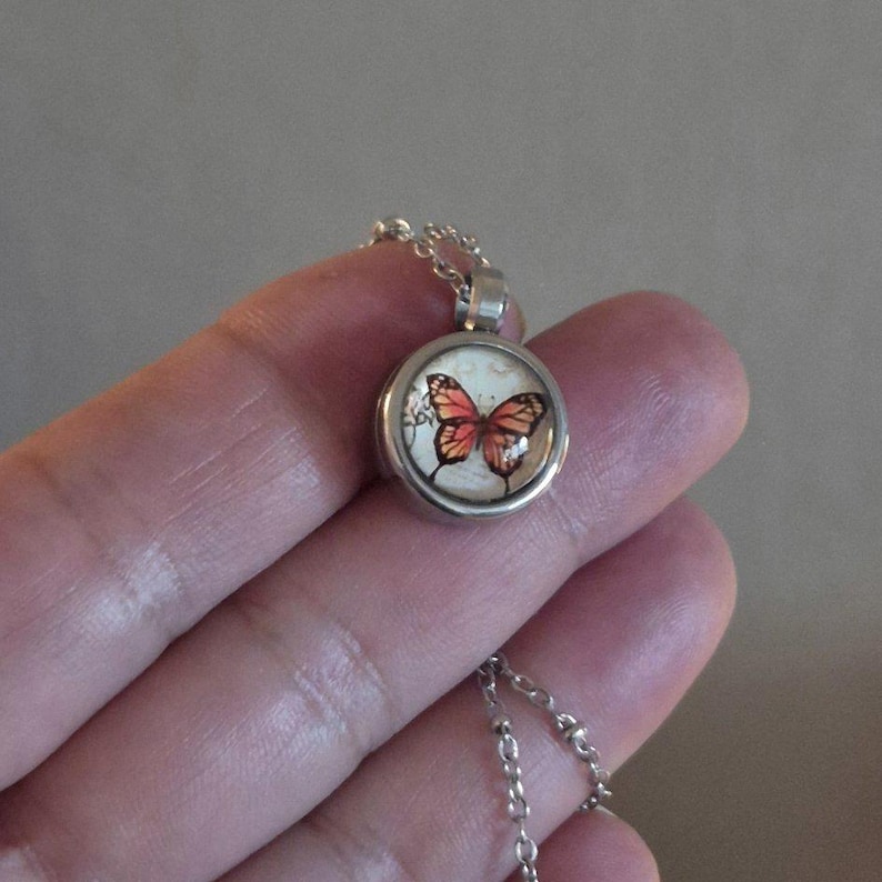 Puede incluir: Un collar con colgante plateado con un dije redondo con un dise&ntilde;o de mariposa. La mariposa tiene alas naranjas y marrones sobre un fondo crema. La cadena del collar es fina y plateada.