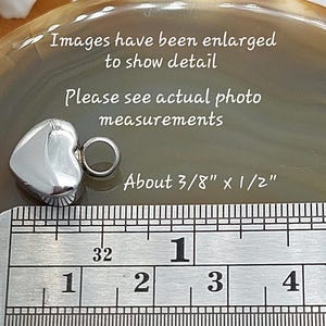 May include: A small, polished silver-toned heart charm with a circular jump ring attached. The charm is positioned next to a metal ruler showing measurements of about 3/8 inch by 1/2 inch. Text on the image reads: Images have been enlarged to show detail, Please see actual photo measurements, About 3/8 inch x 1/2 inch.