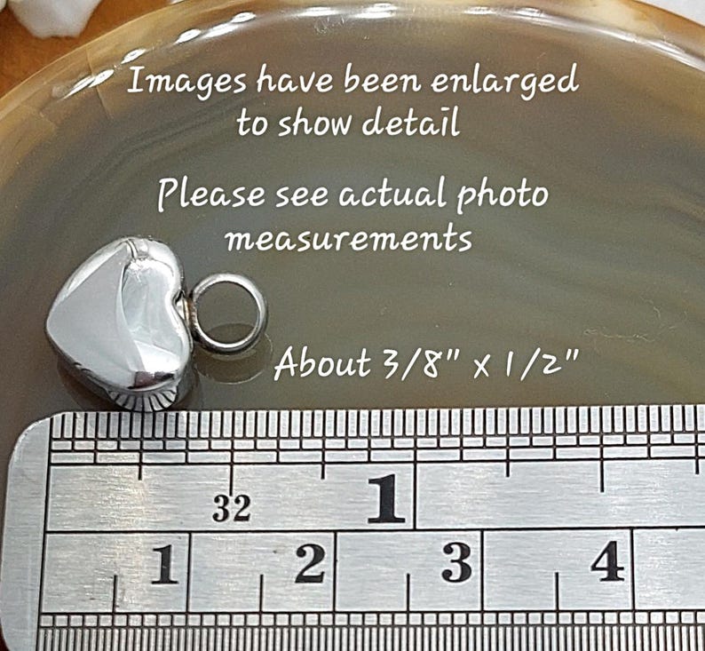 May include: A small, polished silver-toned heart charm with a circular jump ring attached. The charm is positioned next to a metal ruler showing measurements of about 3/8 inch by 1/2 inch. Text on the image reads: Images have been enlarged to show detail, Please see actual photo measurements, About 3/8 inch x 1/2 inch.