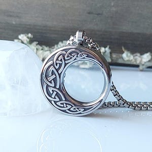 May include: A silver-colored pendant necklace with a circular, Celtic knot design. The pendant has a clear glass center and hangs from a matching chain. The necklace is displayed on a white surface.