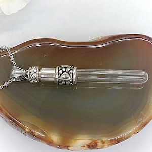 May include: A silver-colored pet urn pendant necklace with a clear glass vial. The pendant features decorative paw prints and scrollwork. The necklace is displayed on a brown and gray surface.