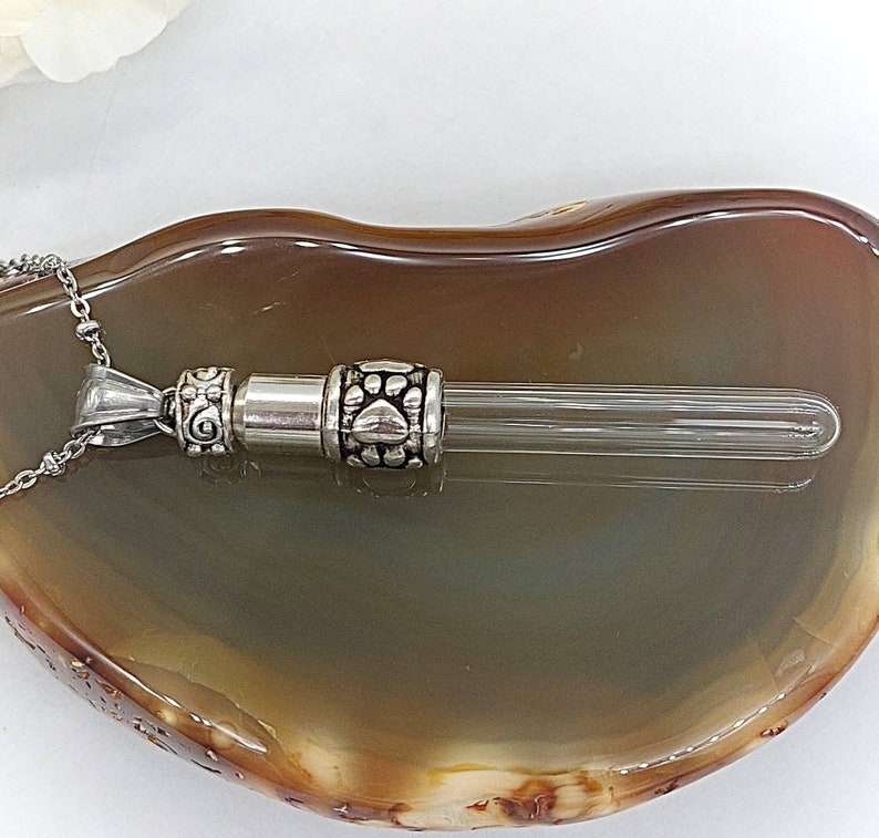 May include: A silver-colored pet urn pendant necklace with a clear glass vial. The pendant features decorative paw prints and scrollwork. The necklace is displayed on a brown and gray surface.