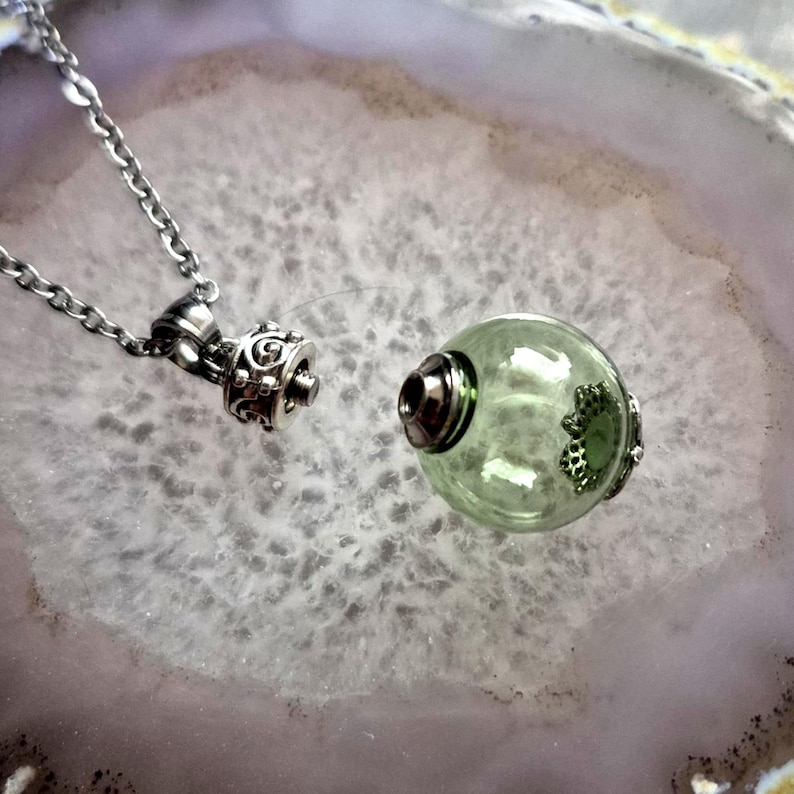 May include: A silver-toned necklace with a decorative pendant and a clear, green-tinted glass orb. The orb has a silver-toned cap and a decorative silver accent. The necklace chain is also silver-toned.
