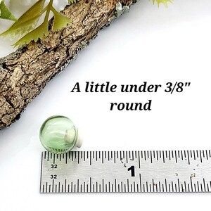 Pu&ograve; includere: Un piccolo globo di vetro rotondo color verde chiaro con un tappo di sughero, posizionato accanto a un righello di metallo. Il testo 'A little under 3/8" round' &egrave; visibile, indicando che il diametro &egrave; inferiore a 0,95 centimetri.