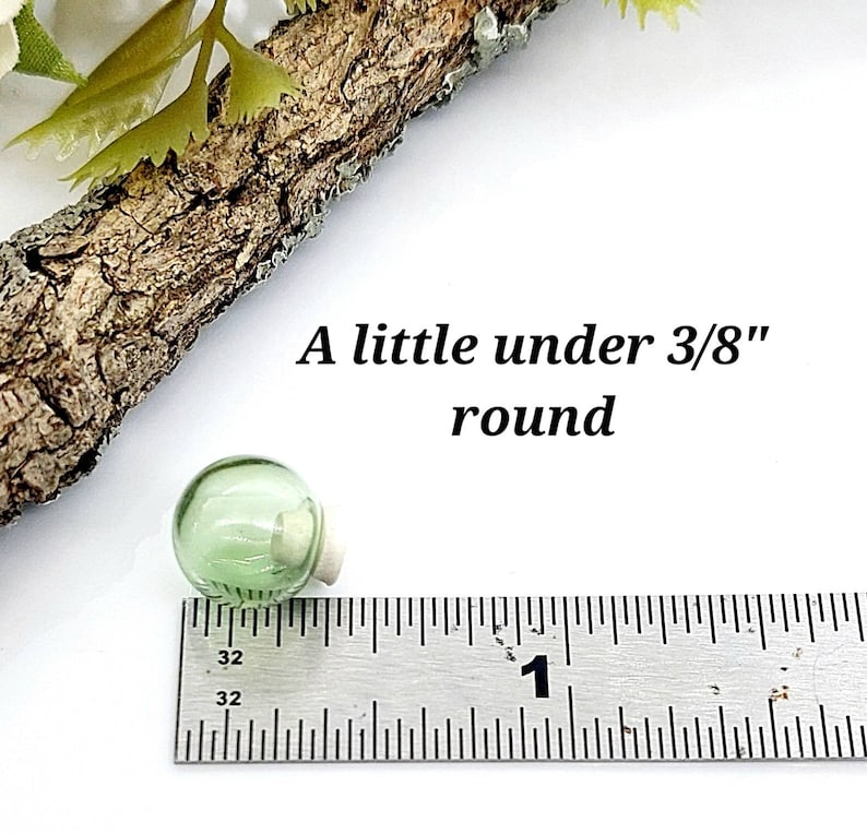 Pu&ograve; includere: Un piccolo globo di vetro rotondo color verde chiaro con un tappo di sughero, posizionato accanto a un righello di metallo. Il testo 'A little under 3/8" round' &egrave; visibile, indicando che il diametro &egrave; inferiore a 0,95 centimetri.