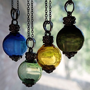 May include: Four glass orb pendants in shades of blue, green, and yellow hang from dark metal chains. Each orb has a decorative metal cap. The pendants are suspended against a blurred background, highlighting the colorful glass.