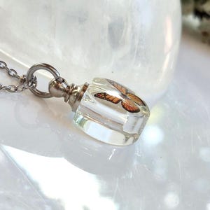 May include: A clear resin pendant shaped like a small bottle, containing an orange and black monarch butterfly wing pattern. The pendant is attached to a silver-toned metal cap and a thin silver-toned chain, creating a necklace piece.