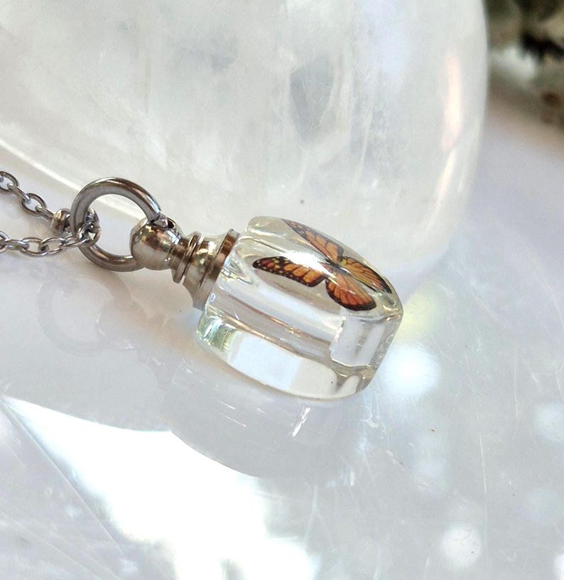 May include: A clear resin pendant shaped like a small bottle, containing an orange and black monarch butterfly wing pattern. The pendant is attached to a silver-toned metal cap and a thin silver-toned chain, creating a necklace piece.