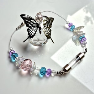 May include: A suncatcher featuring a faceted clear crystal sphere topped with a metallic butterfly silhouette. The circular wire frame is adorned with a sequence of pink, blue, and purple beads, iridescent flower-shaped accents, and a clear glass tube.