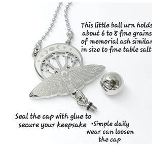 May include: A silver-toned metal pendant necklace featuring a moth and crescent moon design with stars. The pendant includes a small detachable ball urn with a threaded cap. Text states: This little ball urn holds about 6 to 8 fine grains of memorial ash similar in size to fine table salt. Seal the cap with glue to secure your keepsake. Simple daily wear can loosen the cap.
