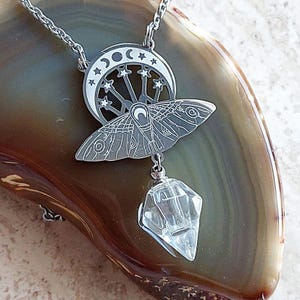 May include: A silver-toned metal pendant necklace featuring a moth design layered over a crescent moon with star cutouts. A clear, faceted, diamond-shaped crystal charm hangs from the bottom of the moth. The pendant is attached to a thin silver chain.