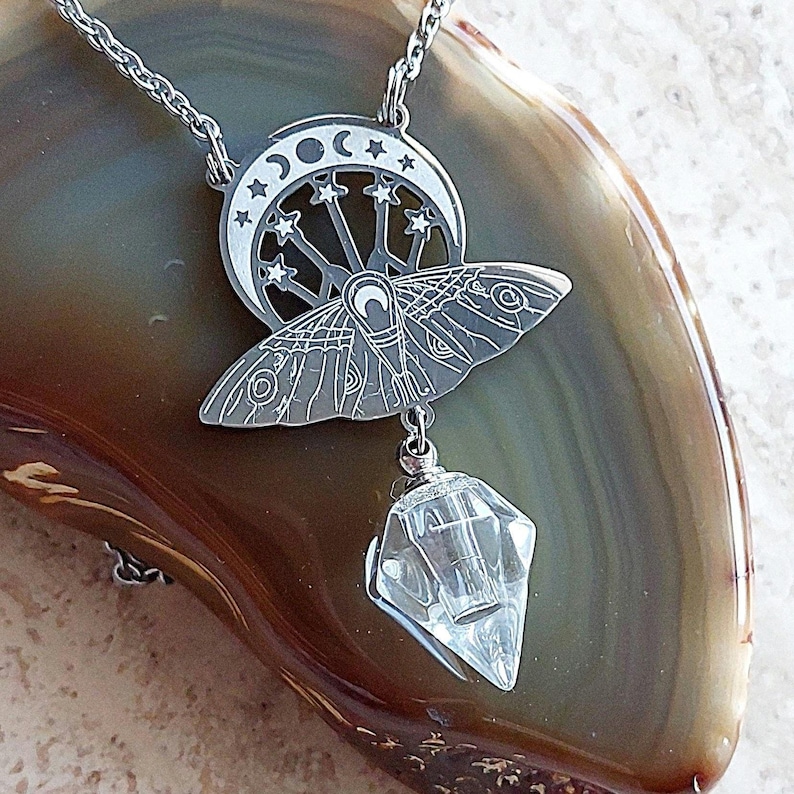 May include: A silver-toned metal pendant necklace featuring a moth design layered over a crescent moon with star cutouts. A clear, faceted, diamond-shaped crystal charm hangs from the bottom of the moth. The pendant is attached to a thin silver chain.