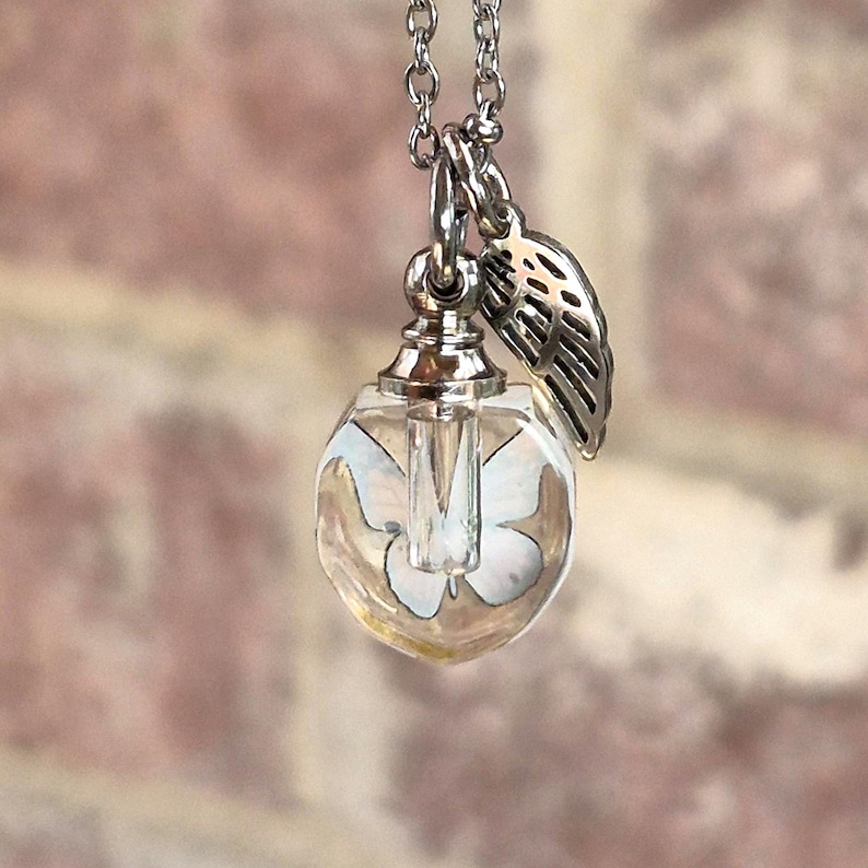 May include: A silver-toned necklace featuring a clear, heart-shaped glass pendant containing a white butterfly graphic. The pendant is topped with a metallic cap and attached to a silver chain, accompanied by a small, detailed silver wing charm.