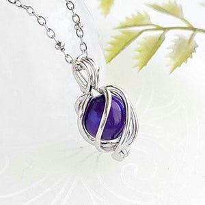 Peut inclure: Un collier en chaîne argent avec un pendentif en pierre précieuse violette enfermé dans une cage en fil d'argent.