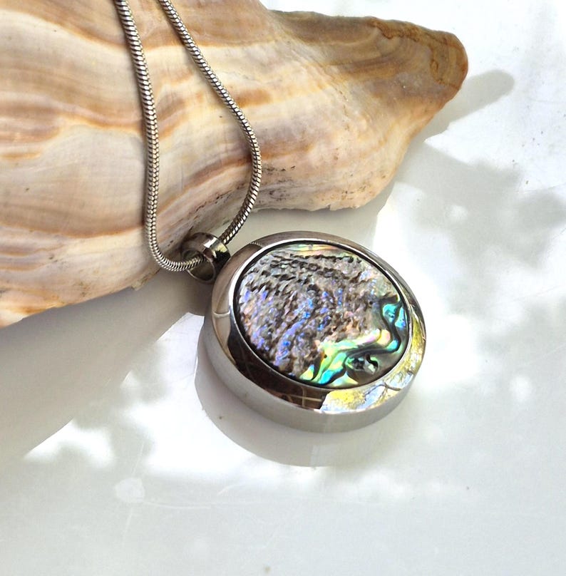 May include: A round silver-toned metal pendant necklace featuring an iridescent abalone shell inlay with swirling blue, green, and purple hues. The pendant hangs from a thin, flexible silver-toned snake chain, showcasing a polished metallic finish.