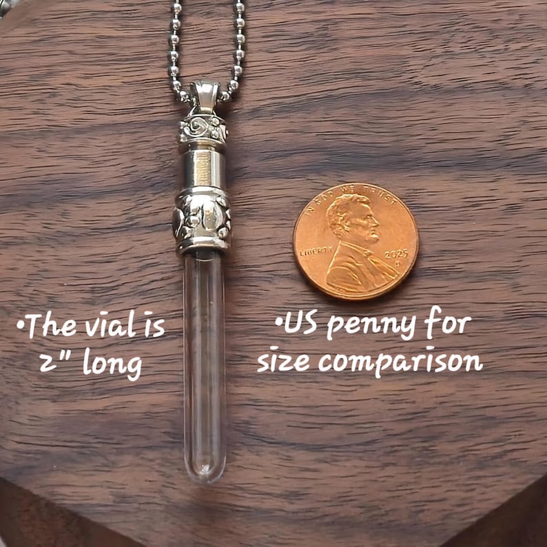 May include: A silver-colored vial pendant with a clear glass tube, suspended from a ball chain. The vial is 2 inches long. A US penny is next to the vial for size comparison. The pendant rests on a dark wood surface.
