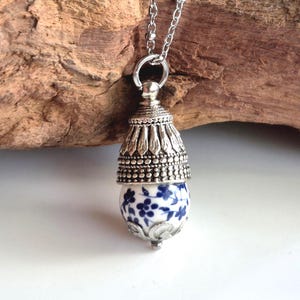 Peut inclure: Un collier pendentif composé d'une perle en céramique blanche à motif floral bleu, insérée dans un capuchon en métal argenté orné de détails perlés et de feuilles, suspendu à une chaîne à maillons en métal argenté assortie.