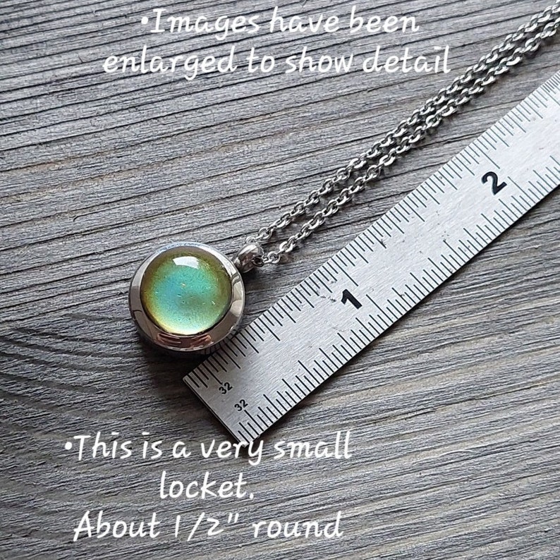 May include: A silver chain necklace with a small round locket. The locket has a green and blue iridescent glass insert. The locket is about 1/2 inch in diameter. The chain is laying on a ruler with the locket at the 1 inch mark.