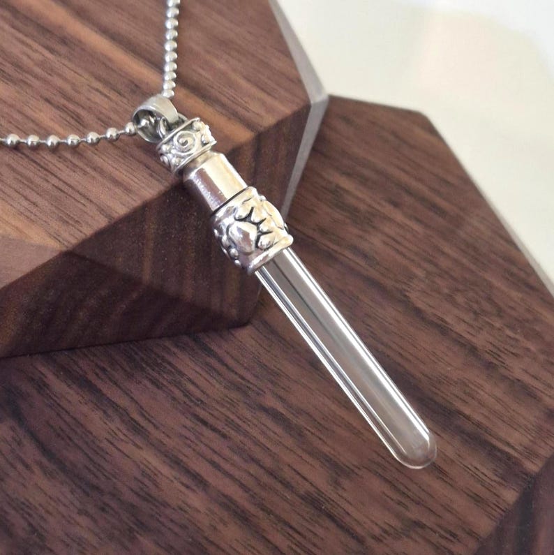 May include: A silver-toned pendant necklace featuring a clear glass vial. The vial is cylindrical with a rounded bottom and is suspended from a decorative silver cap and a ball chain necklace. The pendant rests on a dark wooden surface.