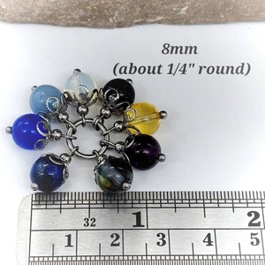 Puede incluir: Un brazalete con siete cuentas redondas de piedras preciosas de 8 mm de di&aacute;metro, en varios colores, incluyendo azul, morado, amarillo y negro. Las cuentas est&aacute;n unidas a una cadena de plata con detalles met&aacute;licos decorativos. El brazalete se mide con una regla, mostrando que las cuentas tienen un di&aacute;metro de aproximadamente 1/4 de pulgada. 8mm (aproximadamente 1/4" de di&aacute;metro)