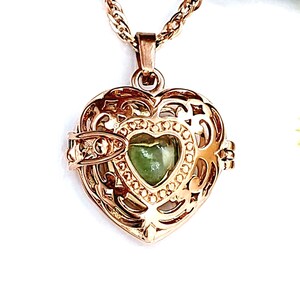 Pu&ograve; includere: Un ciondolo a forma di cuore color oro rosa con un intricato design a gabbia in filigrana metallica. All'interno della gabbia si trova una pietra liscia e lucida a forma di cuore di colore verde chiaro. Il ciondolo pende da una catenina.