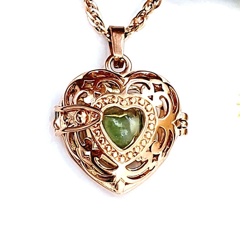 Pu&ograve; includere: Un ciondolo a forma di cuore color oro rosa con un intricato design a gabbia in filigrana metallica. All'interno della gabbia si trova una pietra liscia e lucida a forma di cuore di colore verde chiaro. Il ciondolo pende da una catenina.