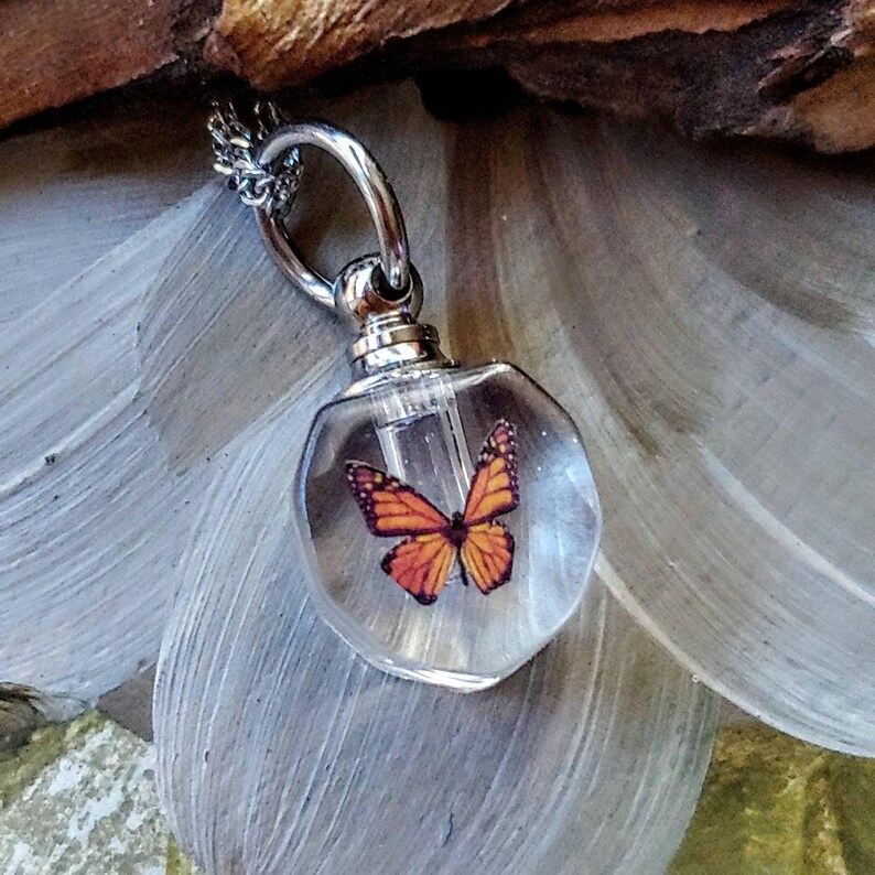 May include: A clear glass pendant shaped like a small vial with a silver-toned metal cap and loop. Inside the transparent glass, a monarch butterfly with orange and black wings is visible. The pendant is attached to a silver-toned metal chain.