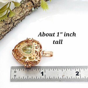 Pu&ograve; includere: Un ciondolo a forma di cuore color oro rosa con intricati intagli in filigrana e una pietra verde chiaro al centro. Il ciondolo presenta un gancio in metallo e misura circa 2,54 cm di altezza, come indicato dal righello presente.