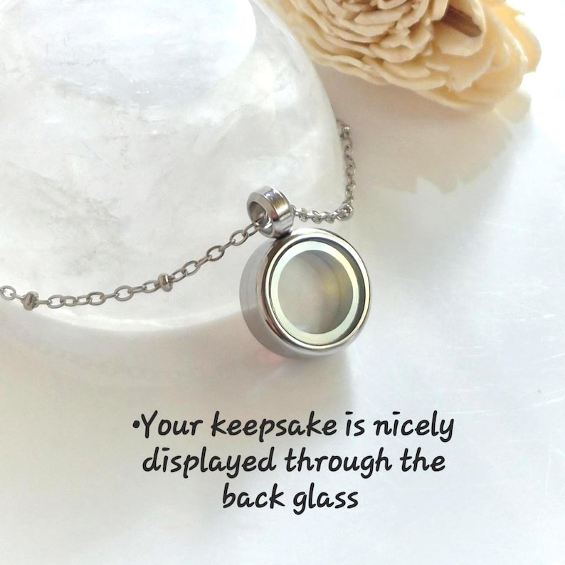 Puede incluir: Un collar con colgante redondo plateado con cristal transparente en la parte delantera y trasera. La cadena es delicada y plateada. El texto de la imagen dice: "Your keepsake is nicely displayed through the back glass."