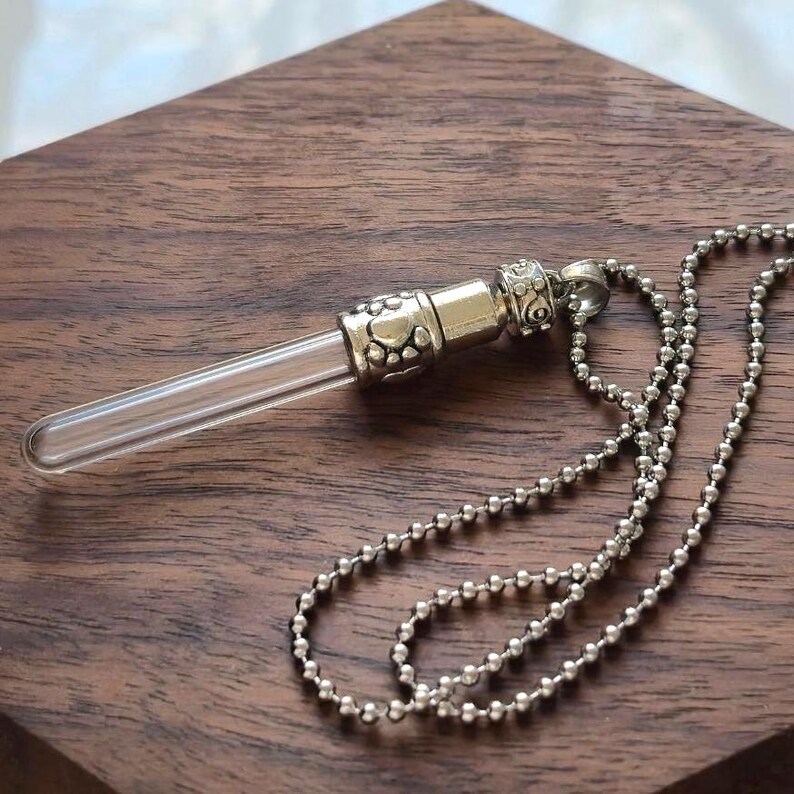 May include: A clear glass vial pendant necklace with a silver-colored metal cap and chain. The vial is cylindrical, and the cap features decorative paw prints and floral designs. The chain is made of small, round beads.