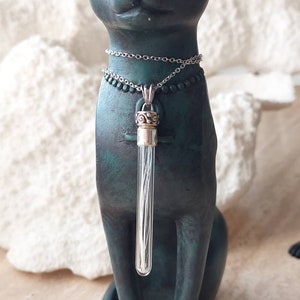 May include: A silver chain necklace with a clear glass vial pendant. The vial is filled with silver thread and has a silver and gold cap. The necklace is worn by a dark green cat figurine.