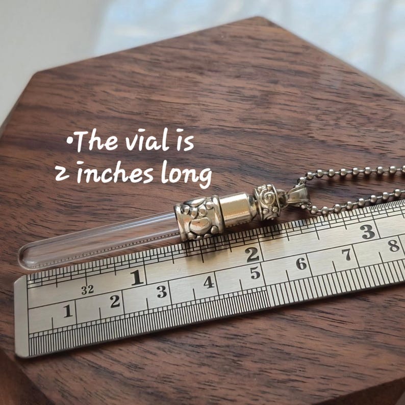 May include: A clear glass vial pendant with silver-colored metal accents and a ball chain. The vial is 2 inches long. The pendant rests on a wooden surface next to a ruler.